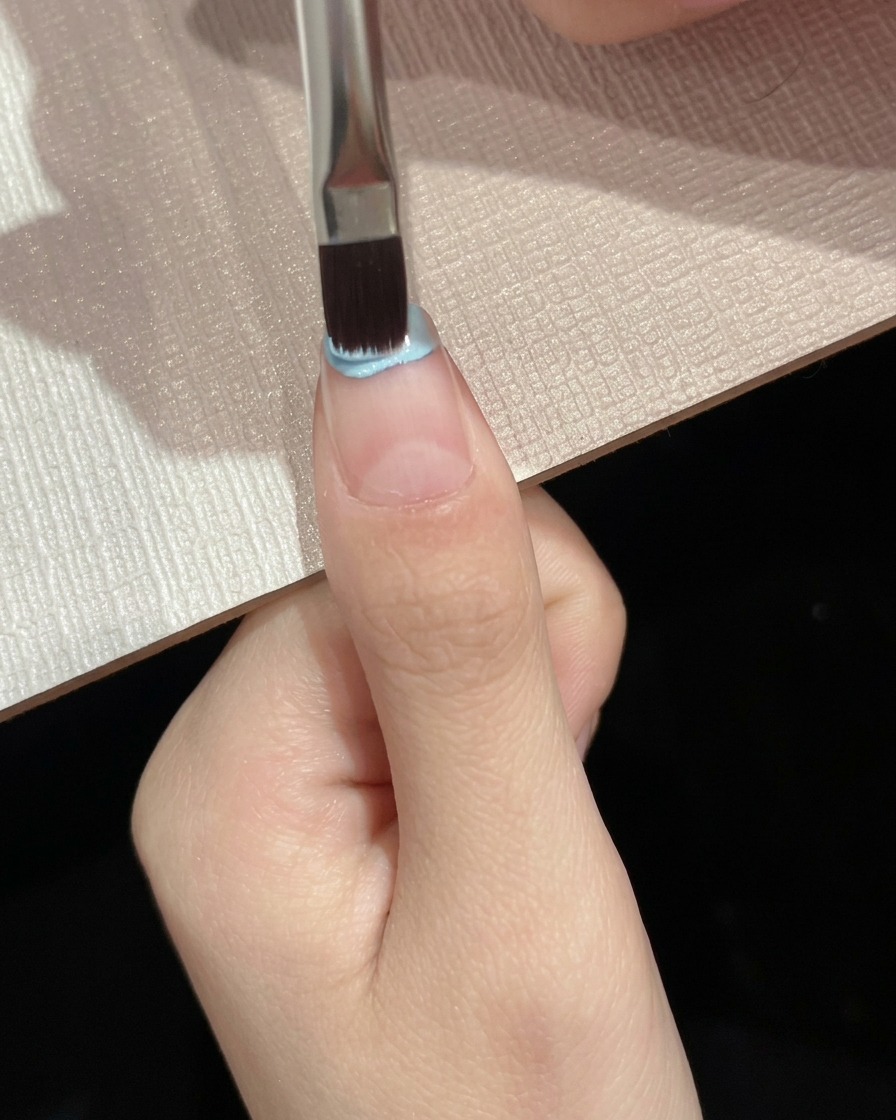 Un pincel plano aplicando esmalte de gel azul claro sobre la mitad superior de la placa de la uña.