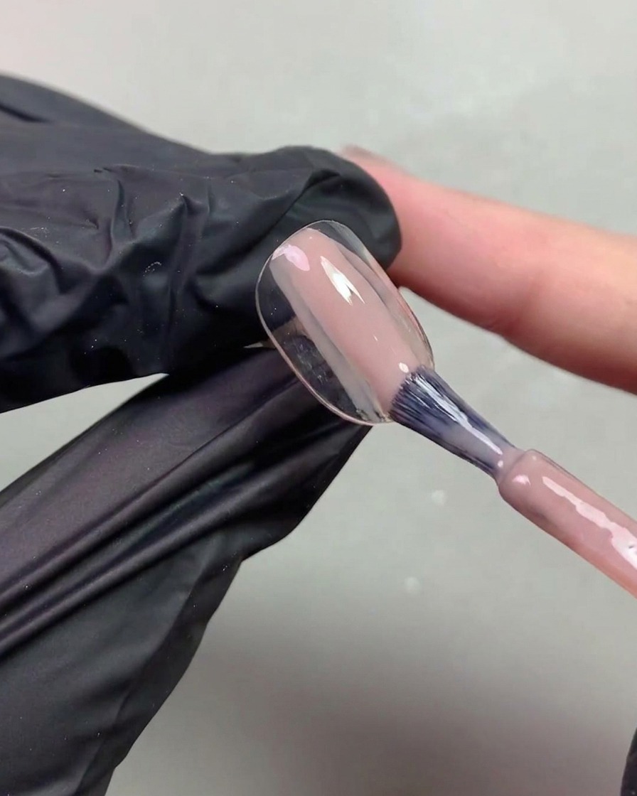 Un primer plano de una mano con guante negro usando un pincel para aplicar un gel traslúcido en tono nude anaranjado sobre una punta de uña.
