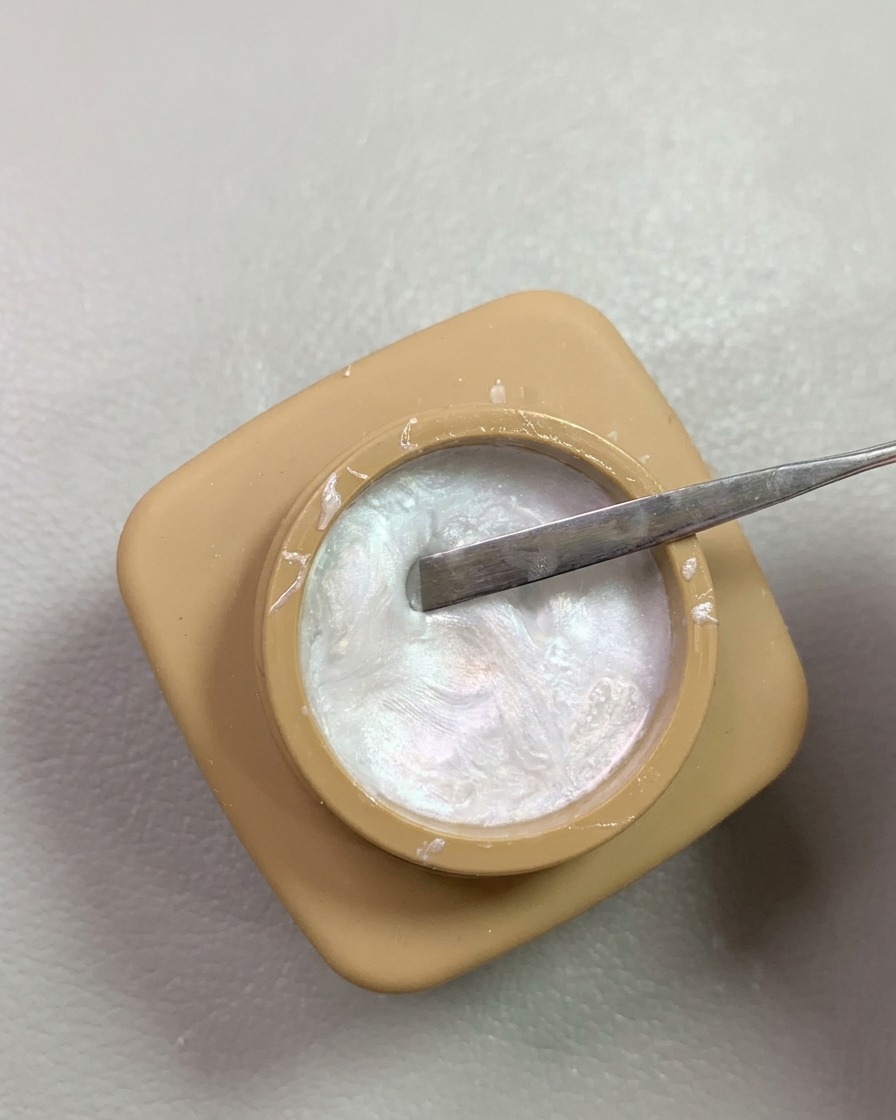 Una herramienta de espátula metálica extrayendo gel de modelado con efecto de perla nacarada brillante de un pequeño tarro color tostado.