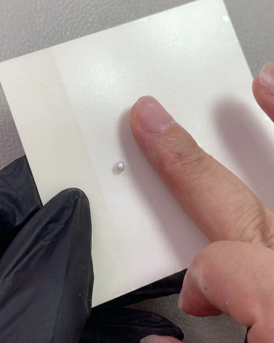 Una mano enguantada señalando una pequeña bola de gel de modelado de perla nacarada colocada sobre un papel de mezcla.