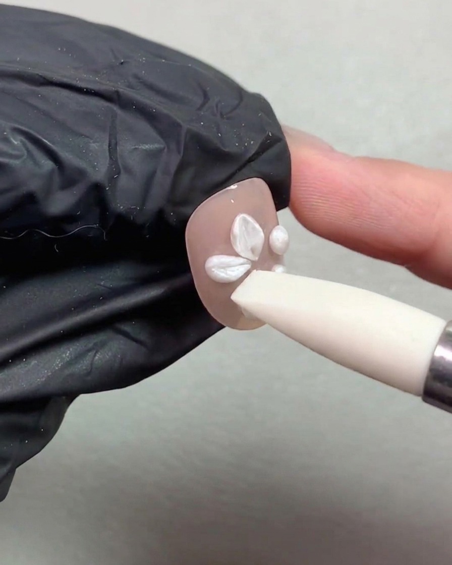 Una herramienta de pincel de silicona presionando y moldeando bolas de gel de perla blanca en pétalos de flores sobre una uña pintada de nude.