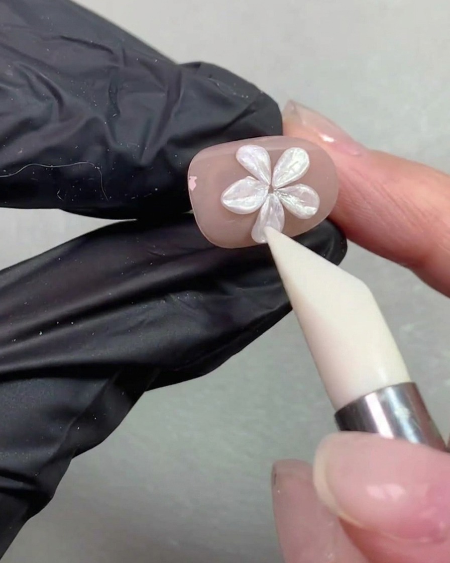 Un pincel de modelado de silicona suavizando y ajustando la forma de los pétalos de una flor de perlas en una uña de color nude.