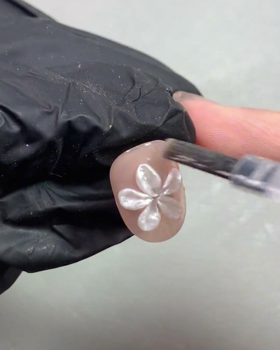 Una mano enguantada colocando una uña corta adornada con una flor de perla blanca en 3D bajo una lámpara de curado UV/LED morada.