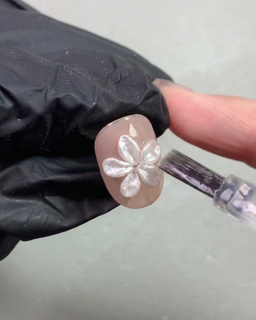 Un pincel para uñas aplicando cuidadosamente una capa fina de capa superior alrededor de una flor de perla 3D curada en una uña.