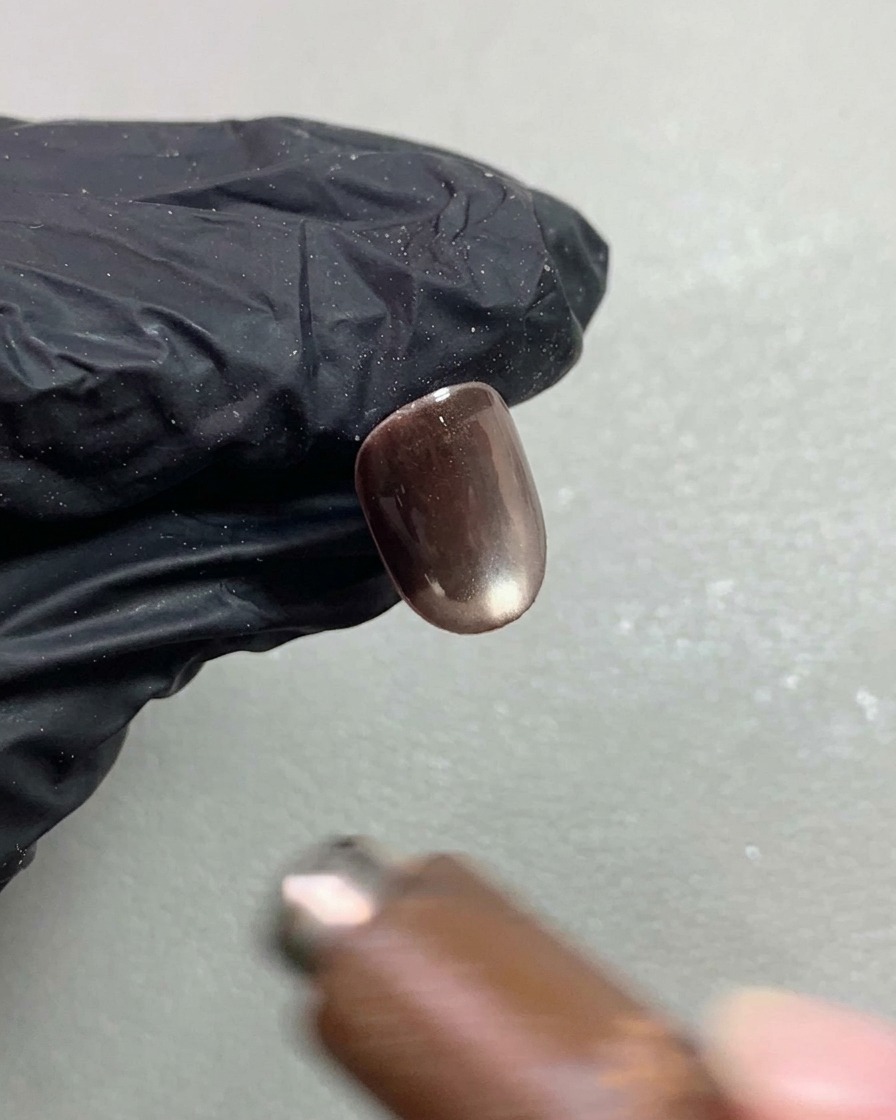 Un imán para uñas flotando sobre una uña corta con gel de ojo de gato marrón oscuro, dando forma al pigmento reflectante en un efecto de esfera brillante en movimiento.