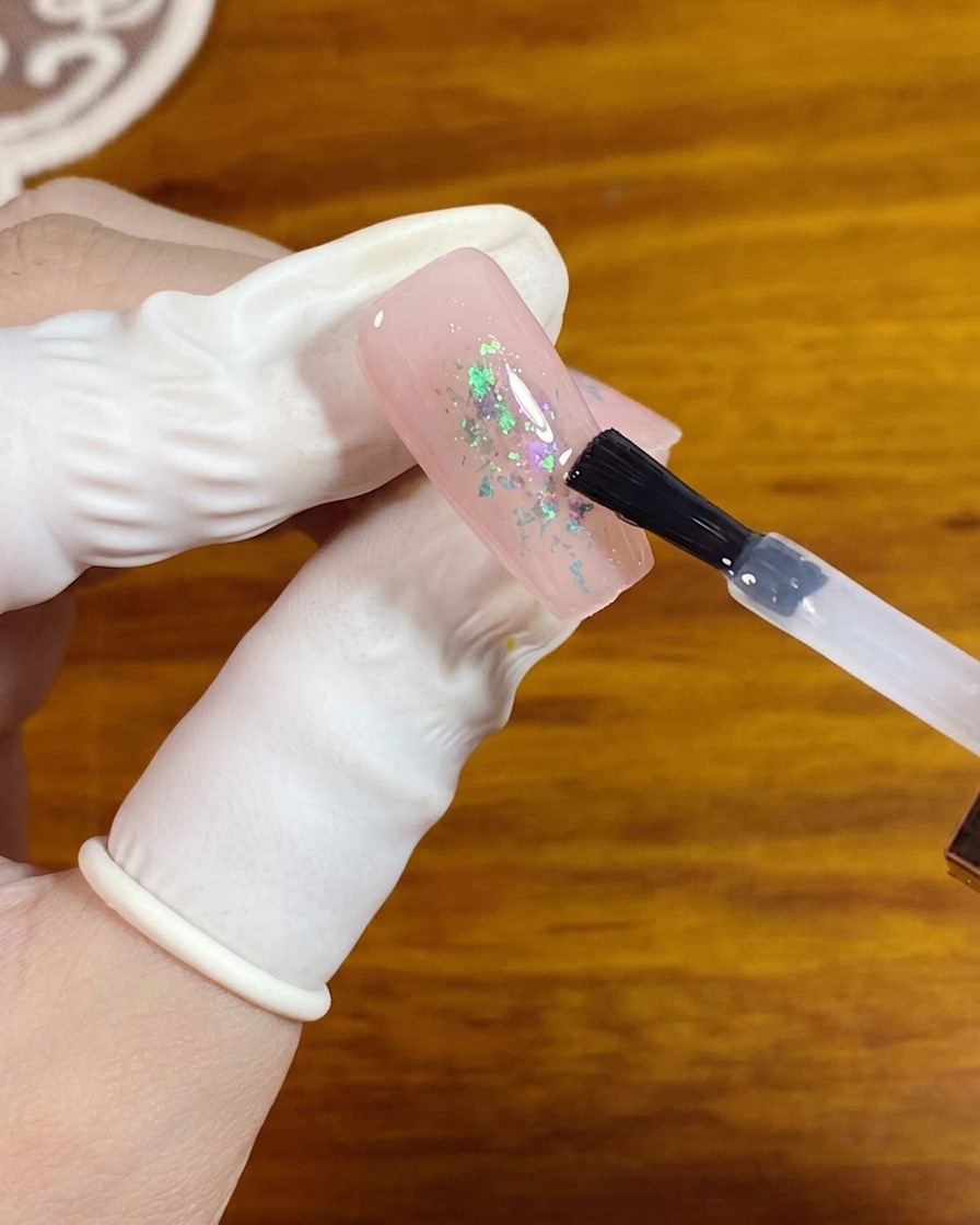 Un pincel de gel aplicando un top coat mate transparente sobre una uña rosa con copos de ópalo en el centro.