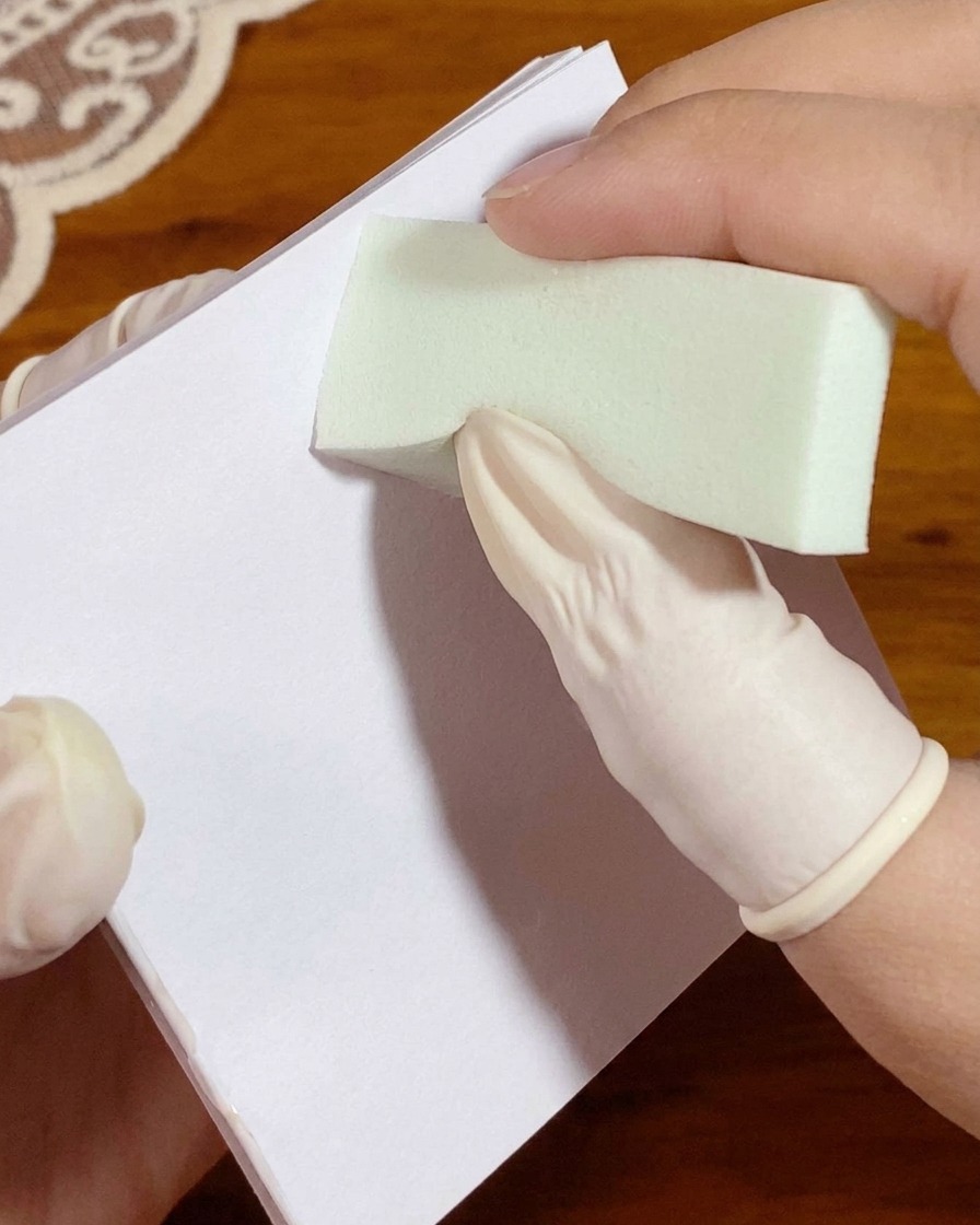 Una mano enguantada presionando una esponja cargada con gel blanco sobre un papel de paleta antes de la aplicación en la uña.