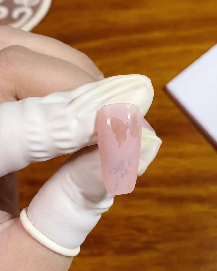 Un primer plano de una mano enguantada sosteniendo una uña postiza que muestra una silueta de mariposa en espacio negativo.