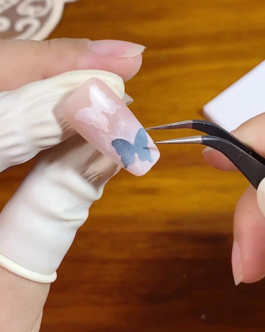 Unas pinzas levantando una pegatina de plantilla de un diseño de uña con degradado rosa.