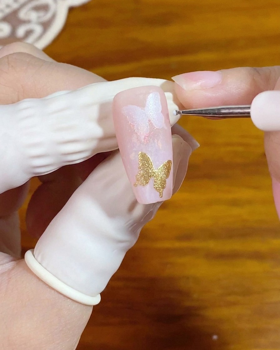 Un pincel de detalle fino aplicando pequeños puntos blancos junto a un elemento de arte de uñas de mariposa dorada.