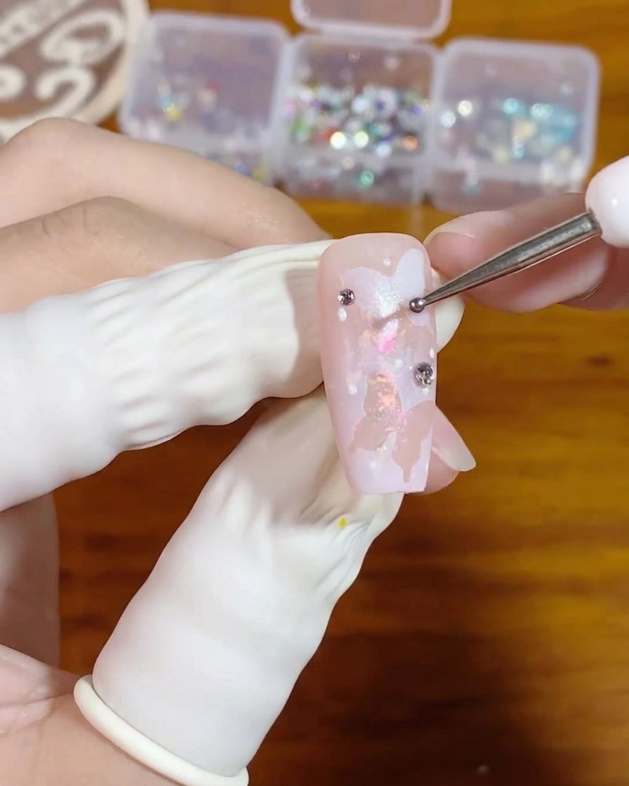 Un primer plano de una mano utilizando una fina herramienta de puntos para colocar una diminuta gota de gel transparente en una uña rosa decorada con mariposas blancas.