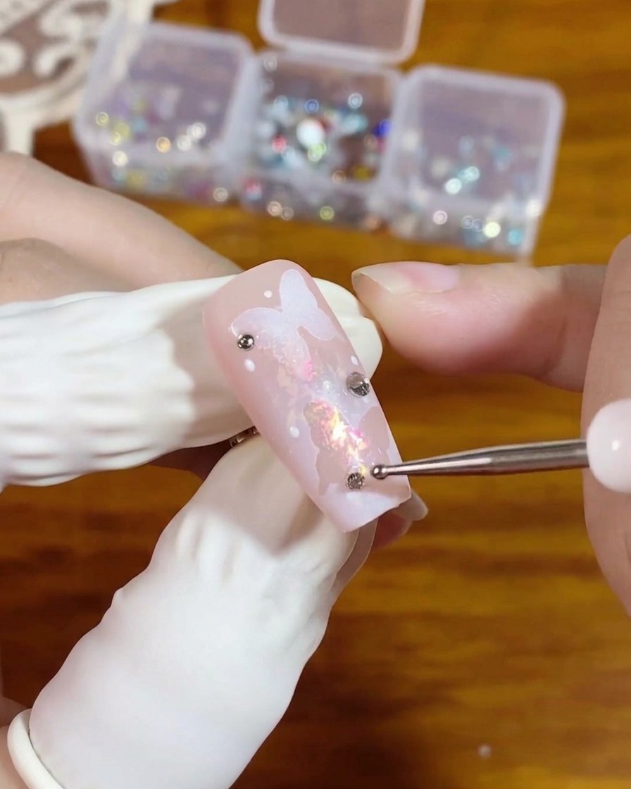 Una herramienta de puntos colocando un pequeño diamante de imitación plateado de parte trasera plana sobre una gota de gel transparente en una uña rosa con diseño de mariposa.