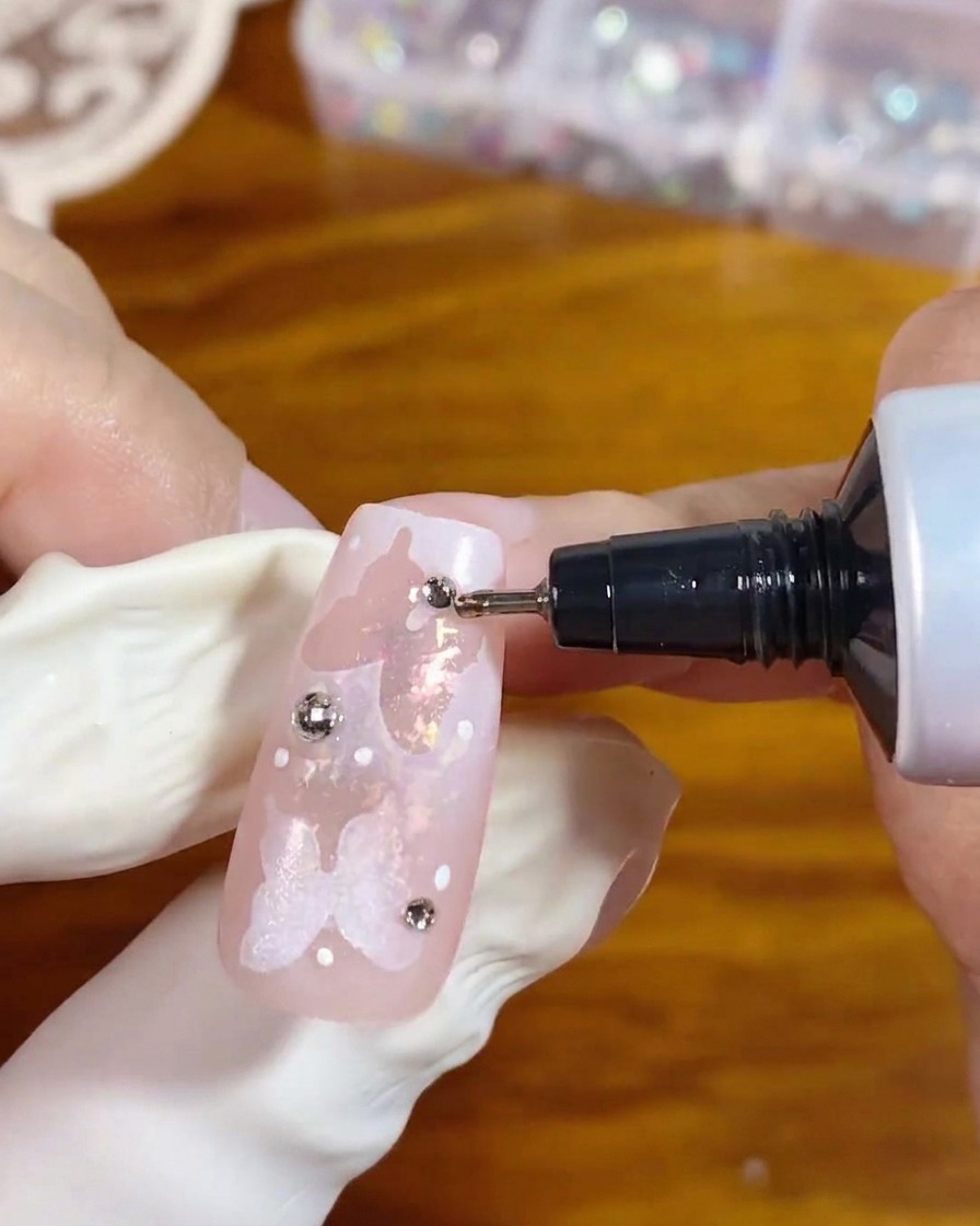 Un tubo aplicador de punta de precisión dispensando gel transparente alrededor de la base de un diamante de imitación plateado en una uña rosa decorada para asegurarlo en su lugar.