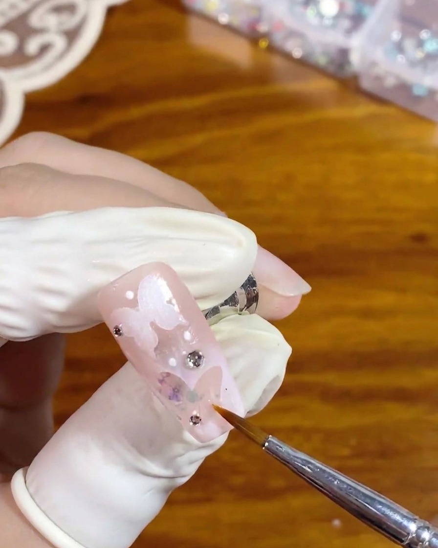 Un pincel delineador fino aplicando cuidadosamente un top coat transparente y brillante alrededor de un diamante plateado en una uña rosa con un diseño de mariposa blanca hueca.