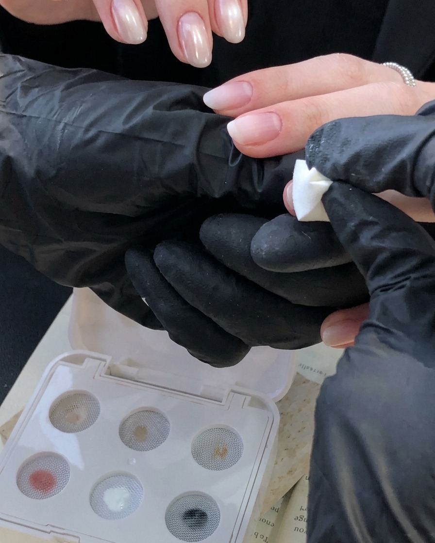 Mano con guante usando una herramienta de sello de esponja blanca para aplicar un degradado blanco marfil en una uña cerca de la cutícula.