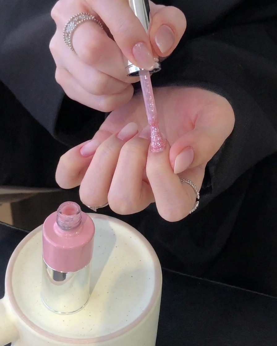 Par de manos sosteniendo un pincel de esmalte rosa sobre un conjunto de uñas manicuradas con un frasco de color rosa sobre un soporte de cerámica.