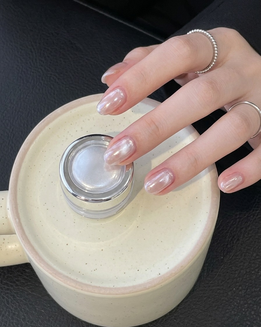 Mano con un anillo sosteniendo un pequeño frasco plateado mientras muestra unas uñas cromadas brillantes terminadas.