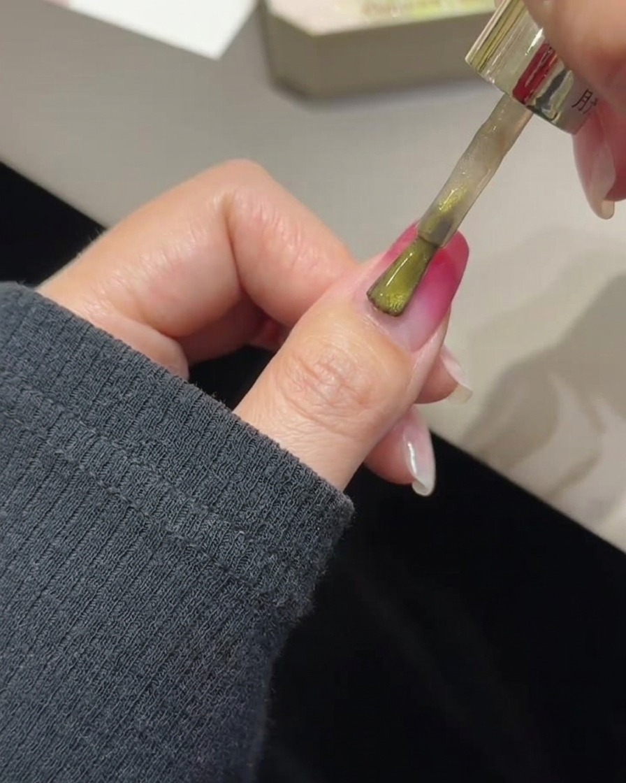 Un pincel aplicando gel cat eye dorado sobre una uña con degradado rosa curado.