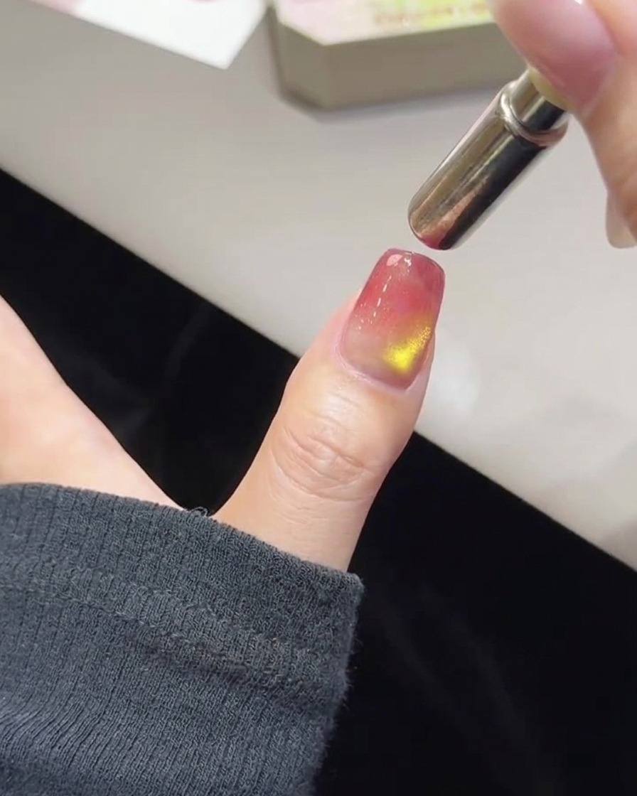 Un primer plano de un pulgar con esmalte rosa y dorado sin curar y una varita magnética cilíndrica suspendida en la punta de la uña.