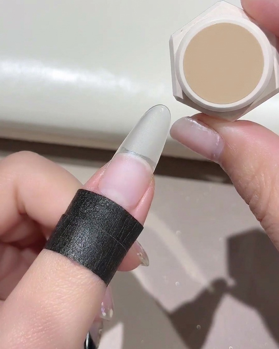 Una pequeña esponja aplicando a toques un color de gel nude sobre una extensión de uña postiza transparente.