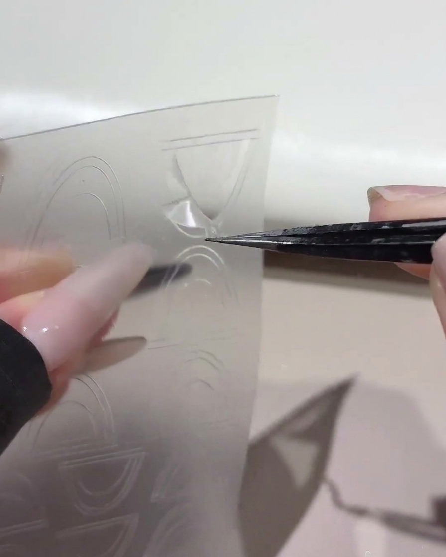 Unas pinzas levantando una pegatina guía curvada para francesa de una lámina de plástico transparente.