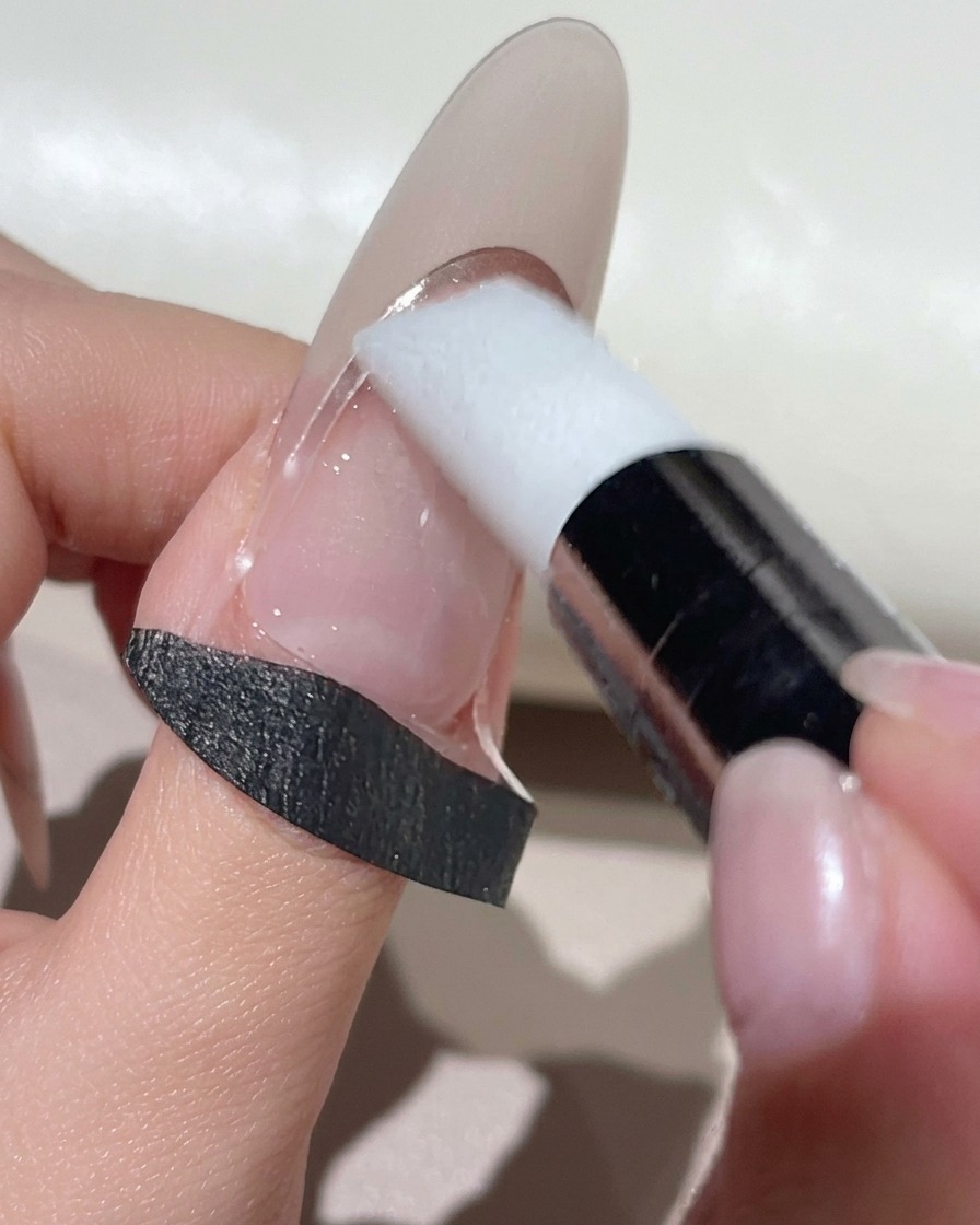Una herramienta de maquillaje de silicona blanca presionando una pegatina guía curvada para francesa sobre una uña en tono natural.