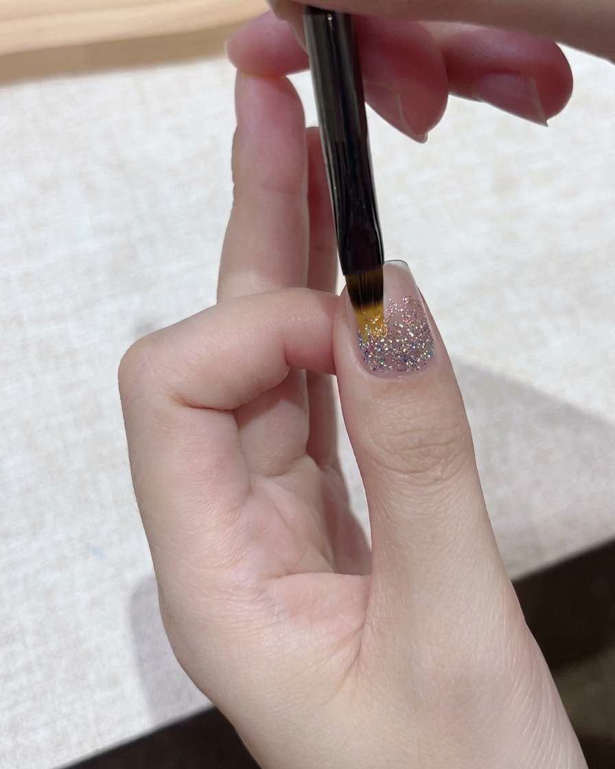 Vista de primer plano de un pincel de detalle fino aplicando una capa de gel transparente sobre una base de uña con glitter brillante.