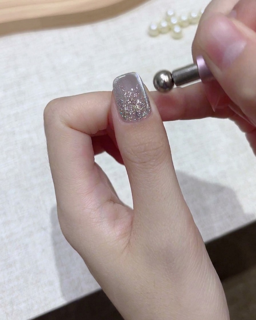 Primer plano de una mano sosteniendo una herramienta magnética esférica sobre una uña de gel con glitter plateado.