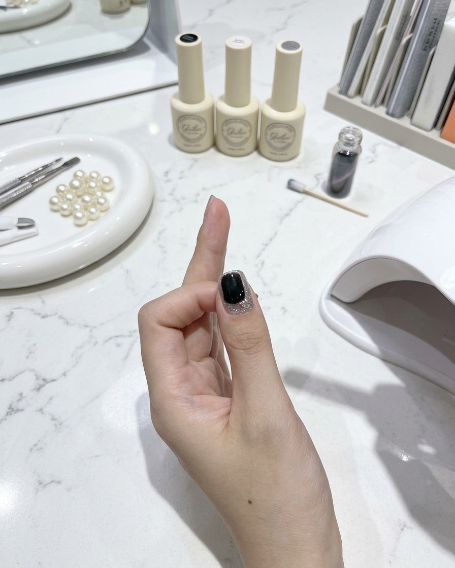 Un pincel de uñas aplicando una capa superior brillante sobre una uña ojo de gato terminada con herramientas de manicura y frascos de esmalte sobre una mesa de mármol blanco en el fondo.