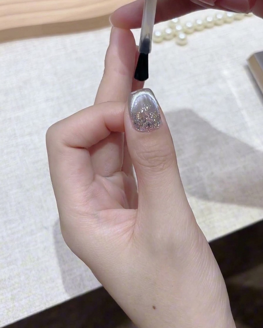 Un pincel aplicando una capa superior clara y brillante sobre una uña de gel magnético plateado terminada.