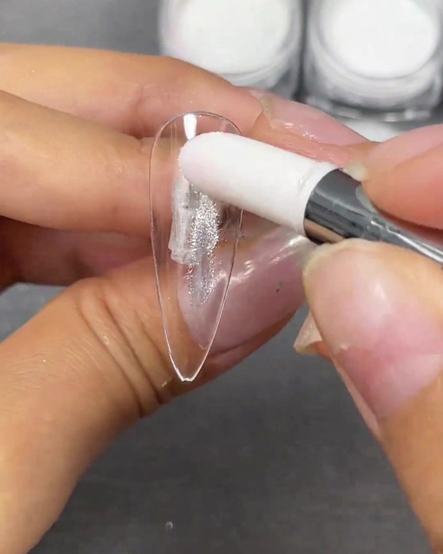 Una mano utilizando un aplicador de esponja blanca para aplicar con toquecitos polvo de chapado en cristal plateado sobre una punta de uña stiletto transparente.