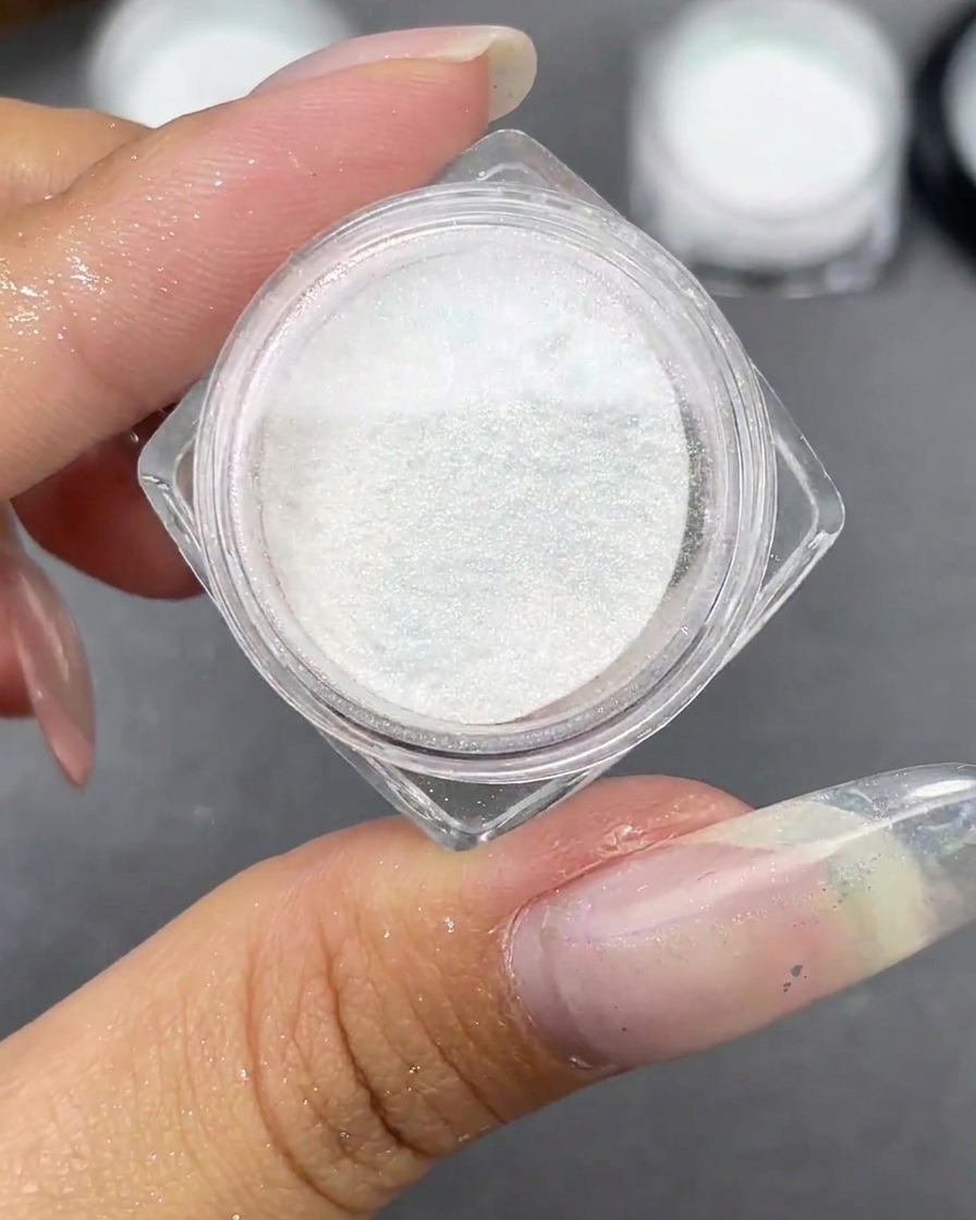 Un frasco pequeño de polvo Moonlight blanco abierto junto a una punta de uña stiletto transparente.