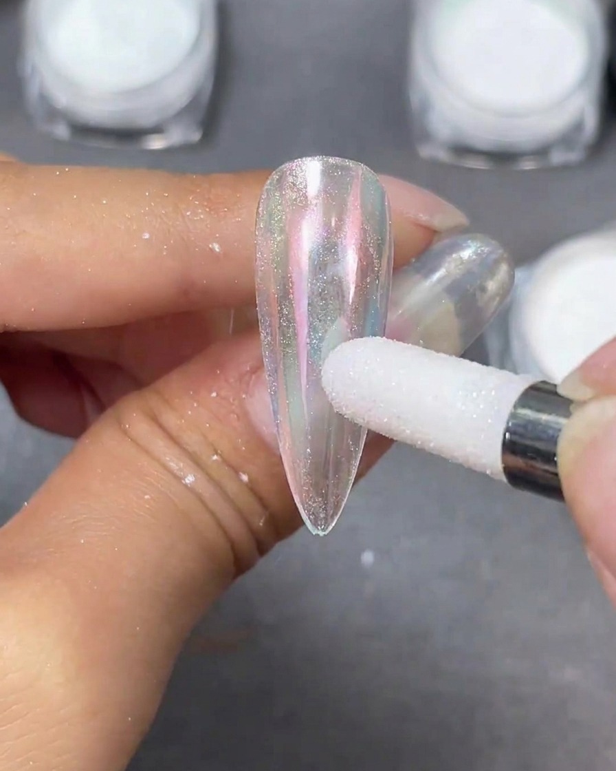 Un aplicador de esponja blanca bruñiendo polvo perlado Moonlight sobre una punta de uña stiletto transparente, creando un cambio iridiscente.