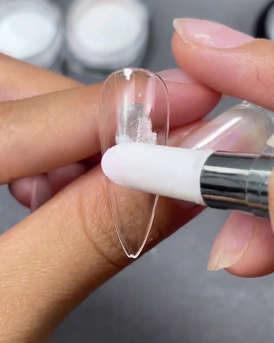 Un aplicador de esponja aplicando toquecitos de polvo Aurora blanco sobre una punta de uña stiletto transparente.