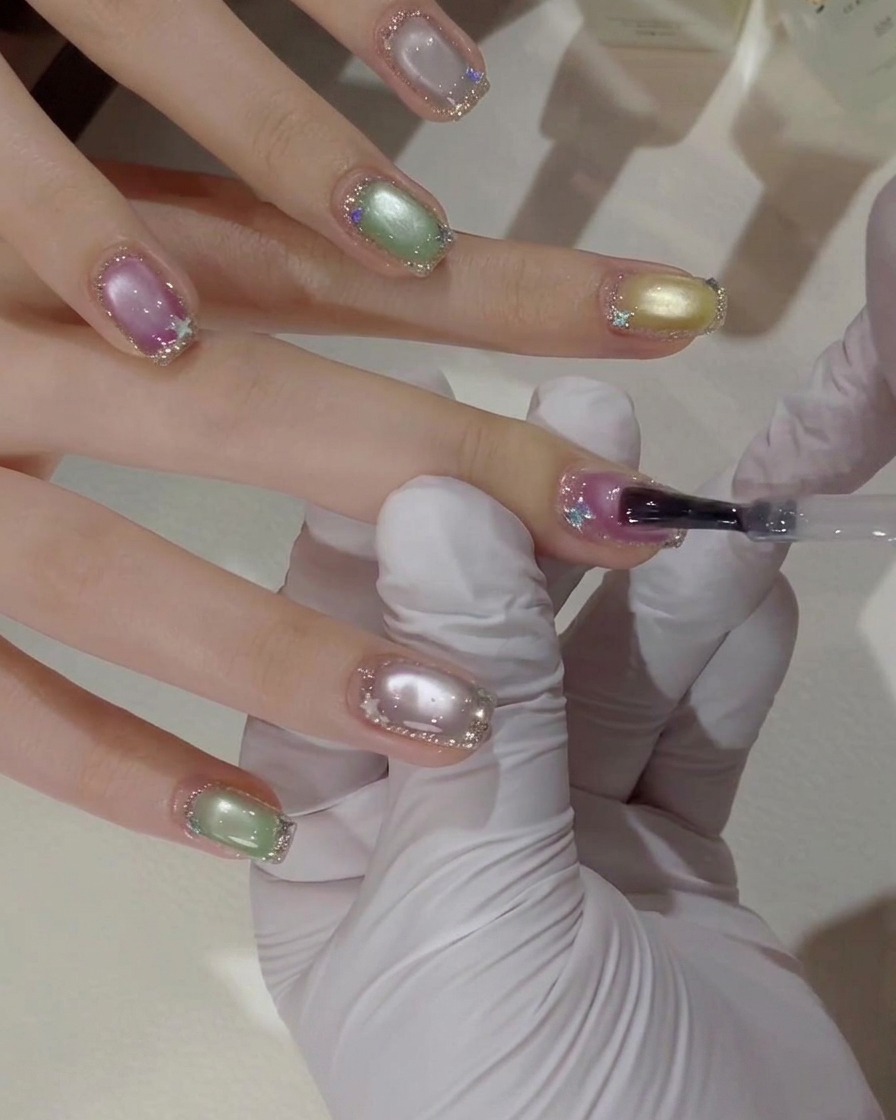 Un primer plano de una mano con guante usando un pincel de gel para aplicar una capa de top coat transparente sobre una uña rosa y morada con efecto ojo de gato y bordes de purpurina plateada.