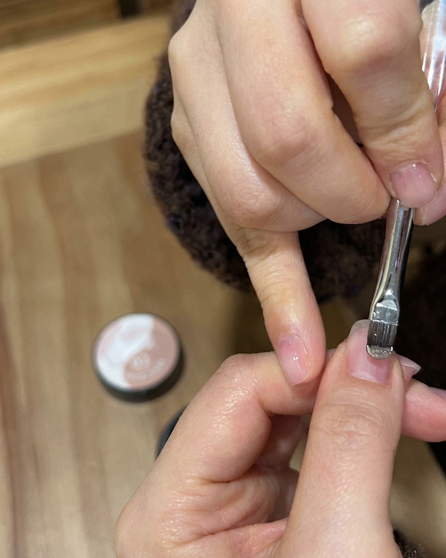 Un primer plano de un pincel de gel aplicando esmalte nude dorado brillante sobre una uña natural.