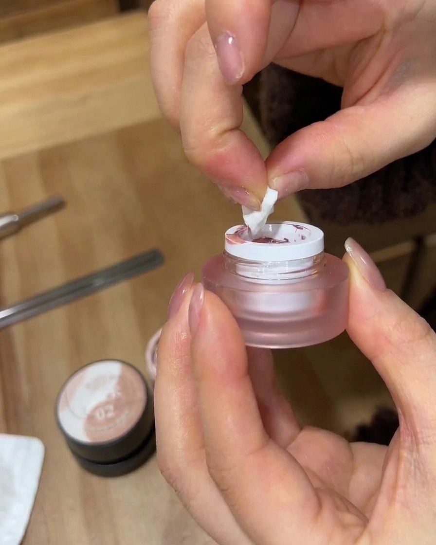 Un primer plano de manos usando un trozo rasgado de esponja de maquillaje blanca para tomar esmalte en gel rosa de un pequeño frasco esmerilado.