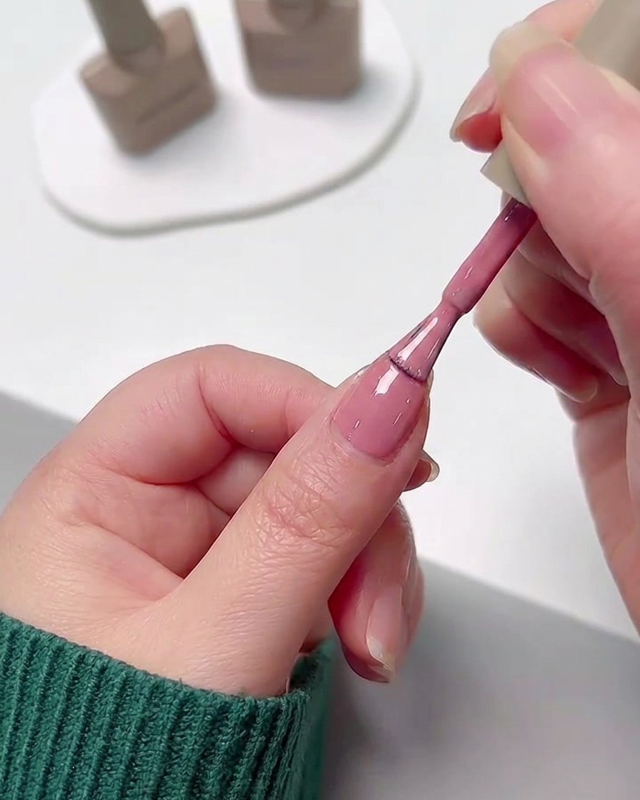 Una mano aplicando una capa suave de esmalte en gel rosa en una uña utilizando el pincel del frasco.