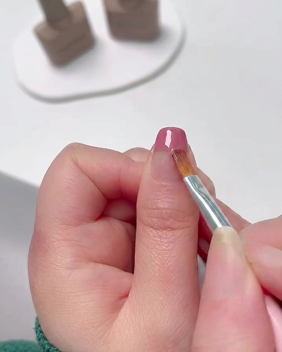 Primer plano de un pincel fino de nail art difuminando esmalte en gel rosa desde la punta de la uña hacia abajo para crear un suave degradado de color.