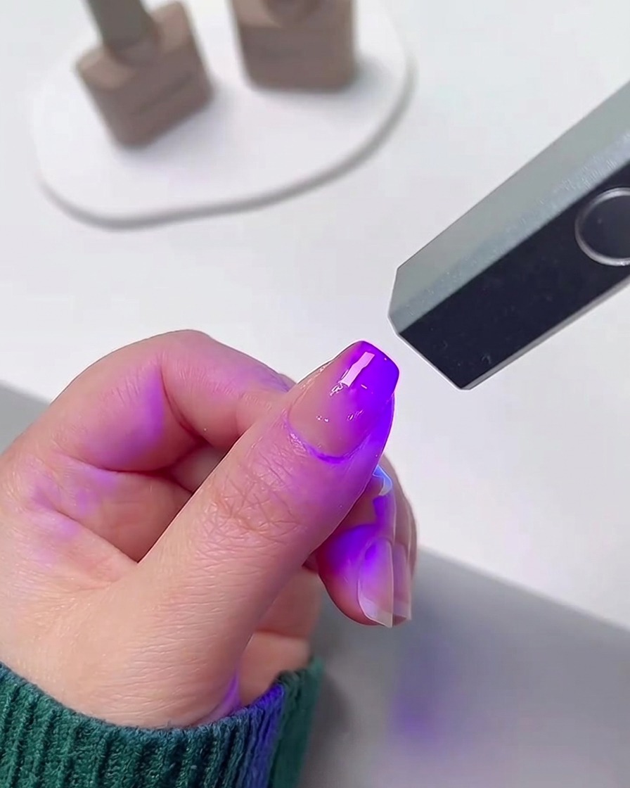 Un dedo posicionado debajo de una lámpara UV/LED pequeña y portátil que emite una luz púrpura brillante para curar el esmalte en gel.