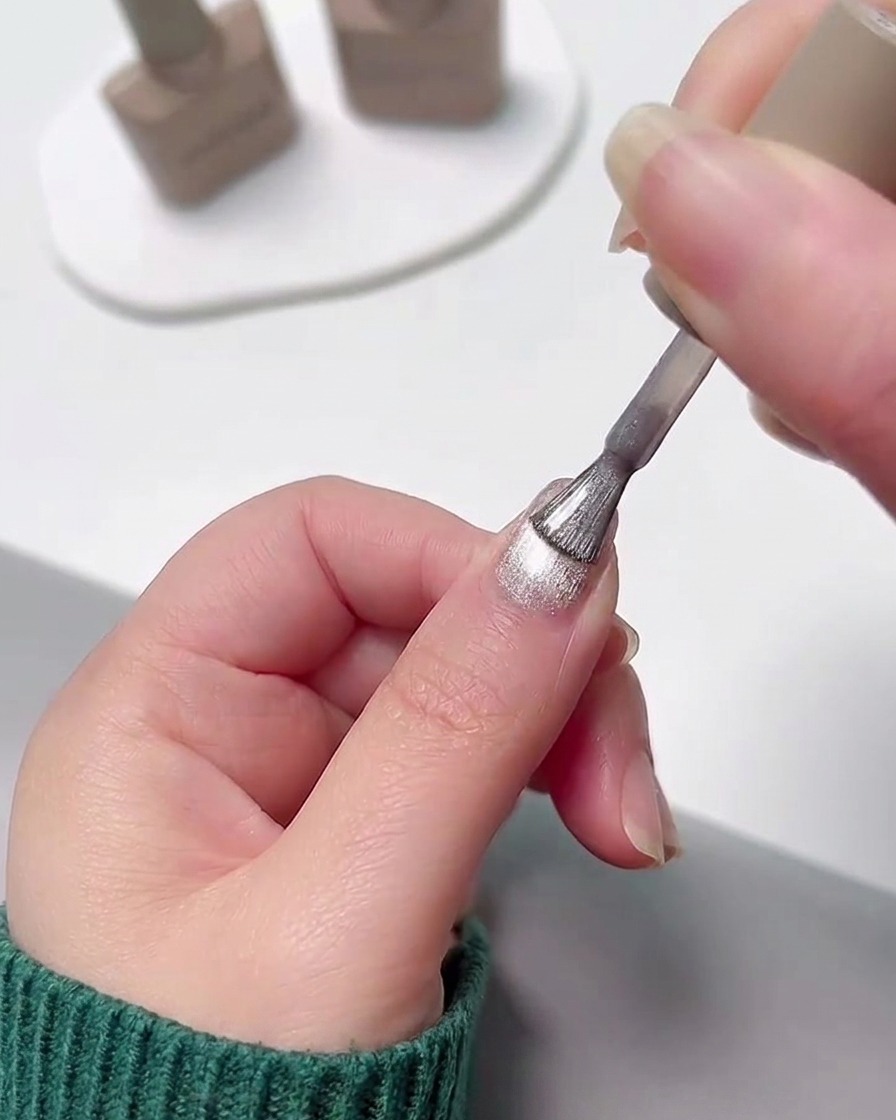 Una mano aplicando una capa de esmalte en gel cat eye plateado brillante sobre una uña con degradado rosa previamente curado.