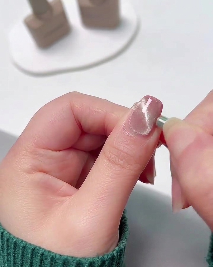 Primer plano de un pequeño imán cilíndrico colocado contra el borde lateral de una uña con gel húmedo y brillante para atraer las partículas metálicas hacia una línea concentrada.