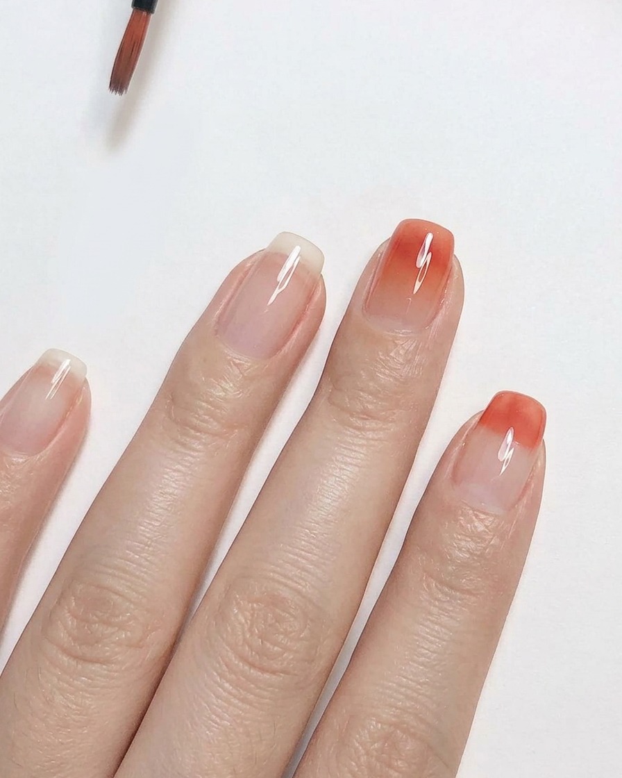 Un pincel de detalle fino difuminando esmalte de gel rosa durazno hacia arriba en una uña transparente para crear un efecto de degradado ombre suave.