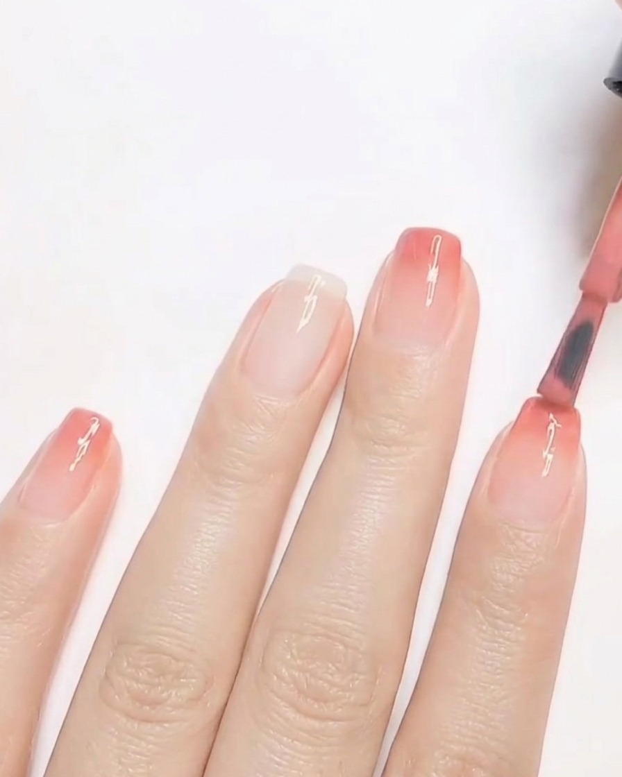 Un pincel de uñas aplicando una segunda capa de esmalte de gel con tono durazno cálido en la punta de una uña para construir un degradado ombre.