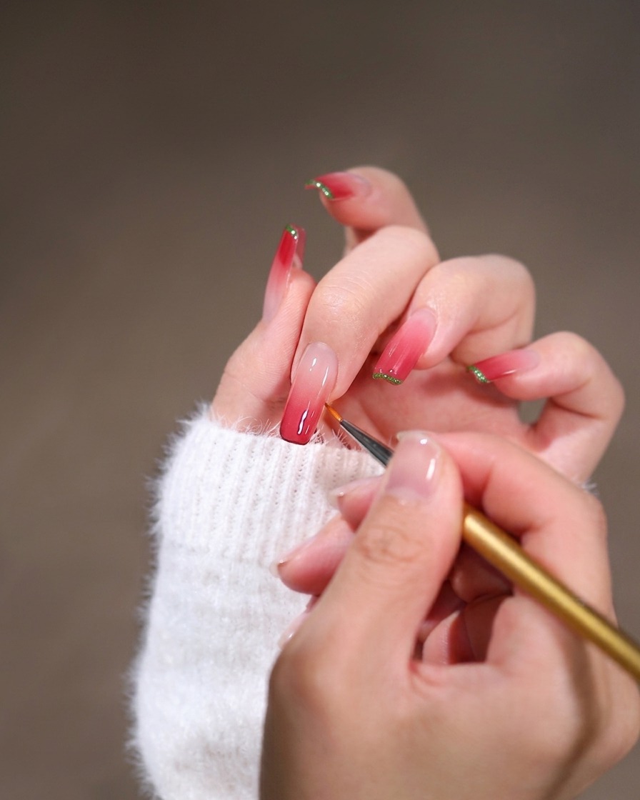 Un primer plano de un pincel liner fino difuminando esmalte en gel rosa a lo largo del borde de la cutícula de una uña cuadrada larga.