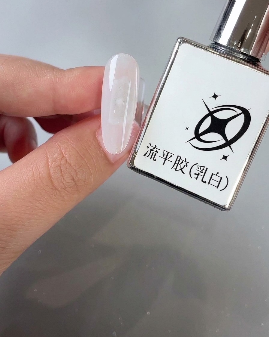 Un pincel aplicando gel lechoso autonivelante sobre la superficie de una uña almendrada junto a su frasco de producto.