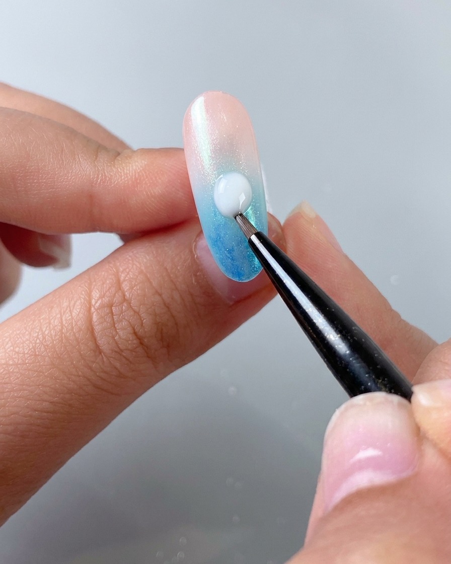 Un pincel fino de nail art aplicando una gota de gel lechoso autonivelante en diagonal a través de una uña con degradado.