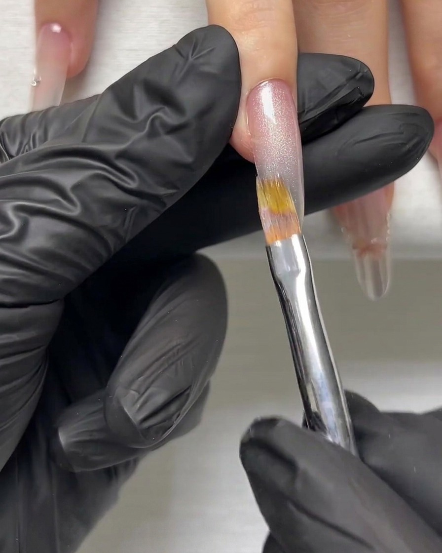 Una mano con guantes usando un pincel fino para suavizar una capa de gel brillante plateado en una uña alargada.