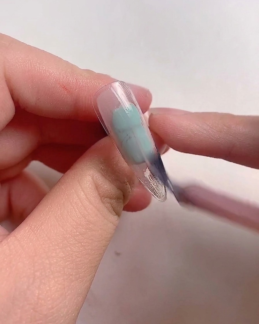 Un pincel aplicando esmalte de gel rosa malva sobre un tip de uña transparente.