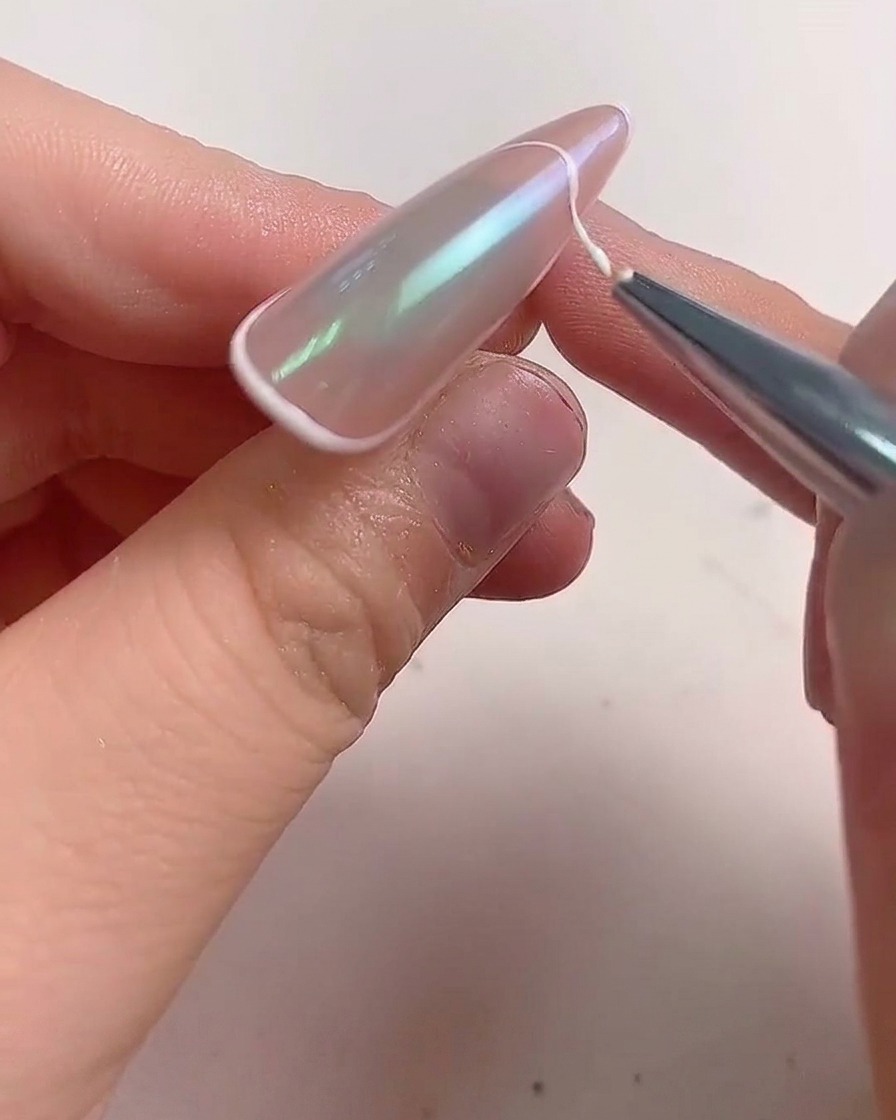 Un pincel delineador fino aplicando una línea de gel blanco a lo largo del borde de una uña almendrada para crear una manicura francesa.